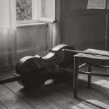 A cello lying on its side on the worn floorboards of an empty music room, its bow resting across a wooden chair, a music stand and a curtained window behind, black and white photograph © The Voice Realm