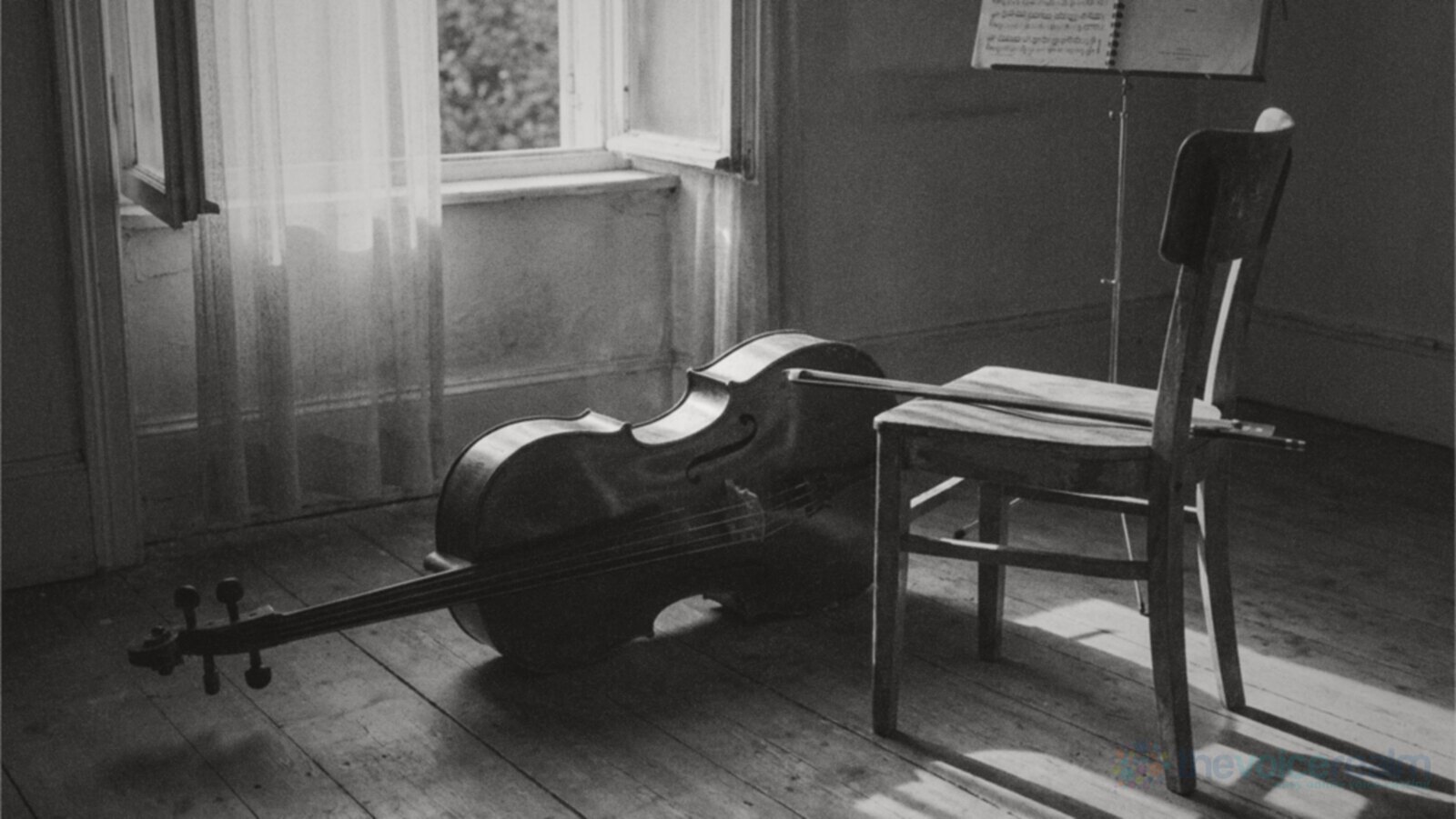 A cello lying on its side on the worn floorboards of an empty music room, its bow resting across a wooden chair, a music stand and a curtained window behind, black and white photograph © The Voice Realm
