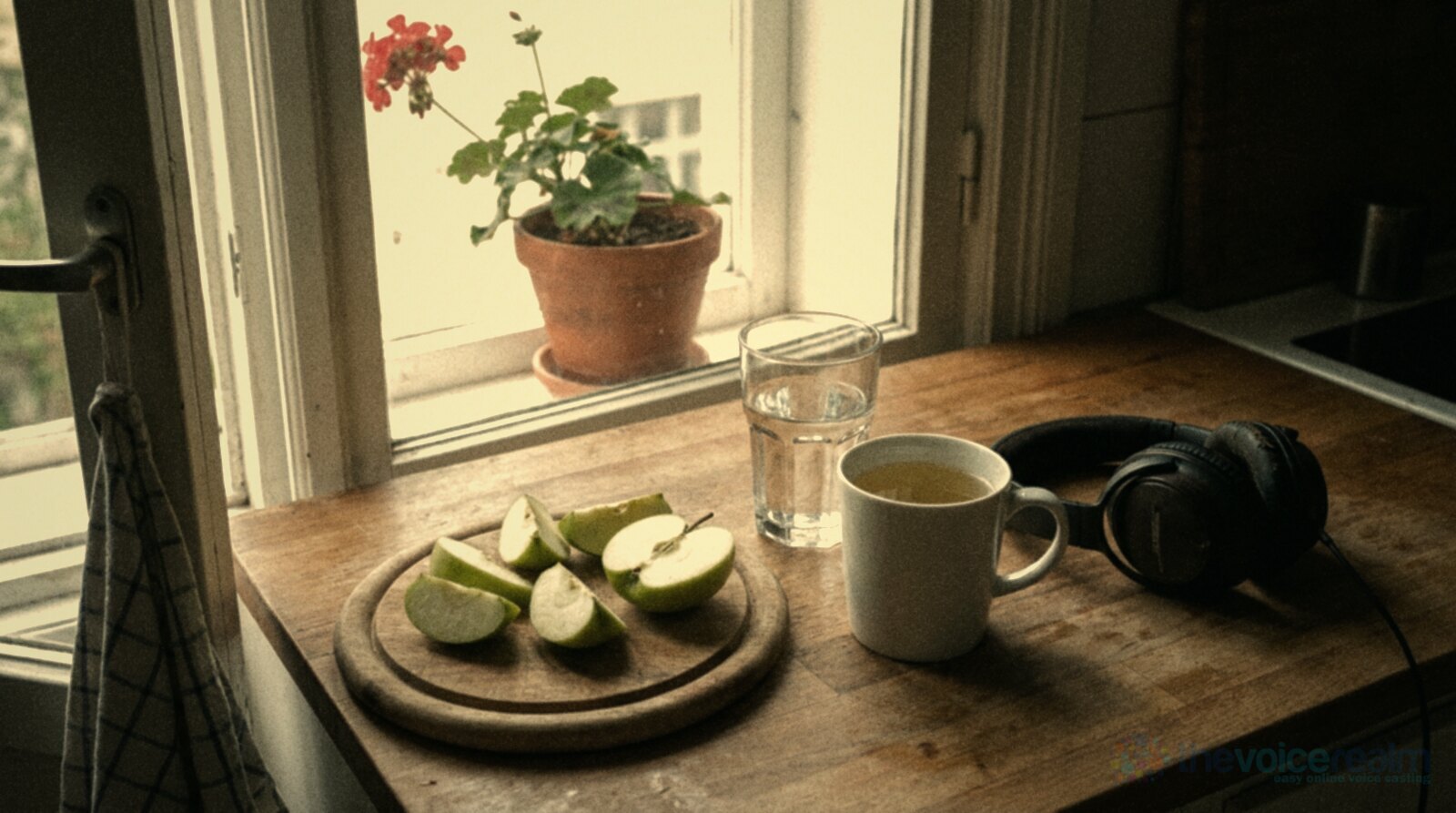 A wooden kitchen counter by a window in morning light: green apple slices on a round wooden board, a glass of water, a white mug of pale herbal tea and a pair of closed-back headphones, a red geranium in a pot on the sill, in soft warm faded film colours © The Voice Realm