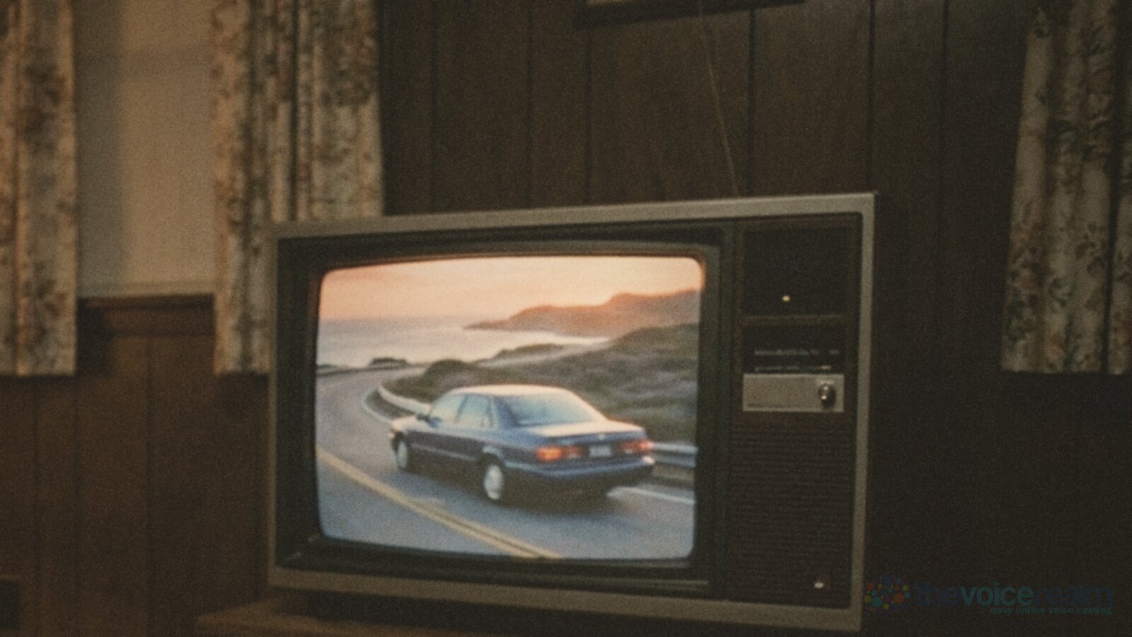 A 1980s wood-panelled living room with floral curtains and a boxy television set showing a dark blue sedan driving a coastal road at sunset © The Voice Realm