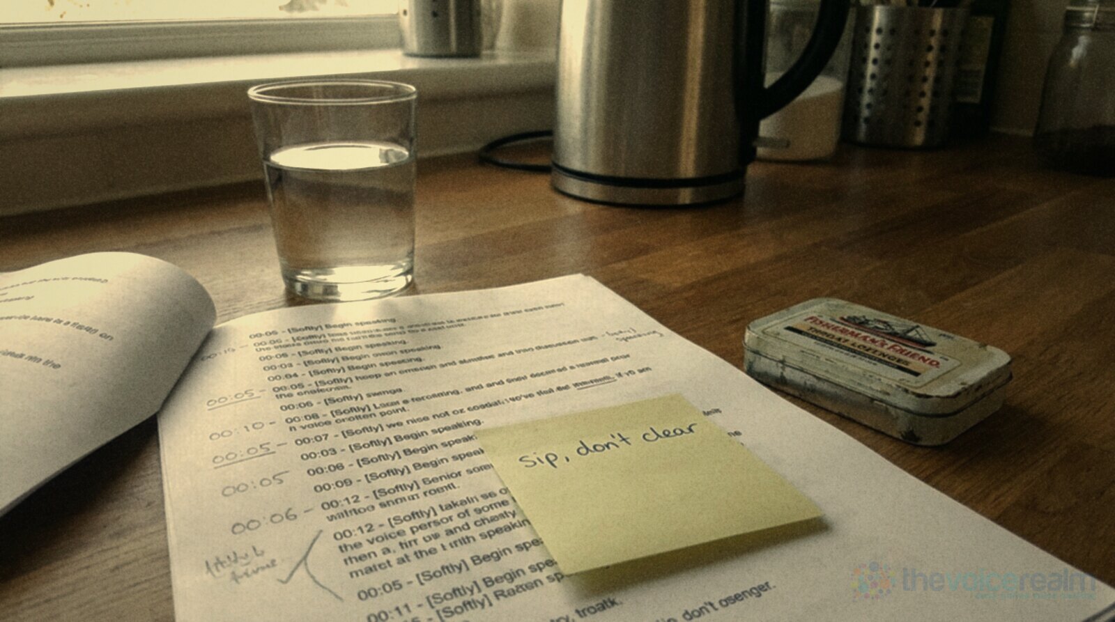A kitchen counter in morning light with a glass of water, a tin of Fisherman's Friend lozenges and a printed script carrying a yellow sticky note that reads Sip, don't clear © The Voice Realm