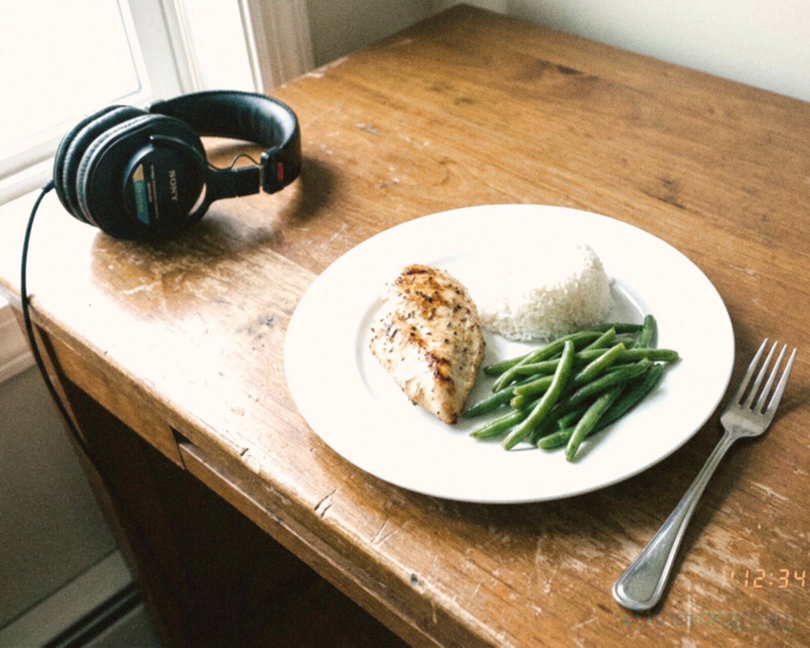 A plain lunch on a white plate on a scuffed wooden desk by a window: grilled chicken, steamed rice and green beans with a fork, studio headphones resting at the edge of the desk, in bright, flat phone photo colours © The Voice Realm