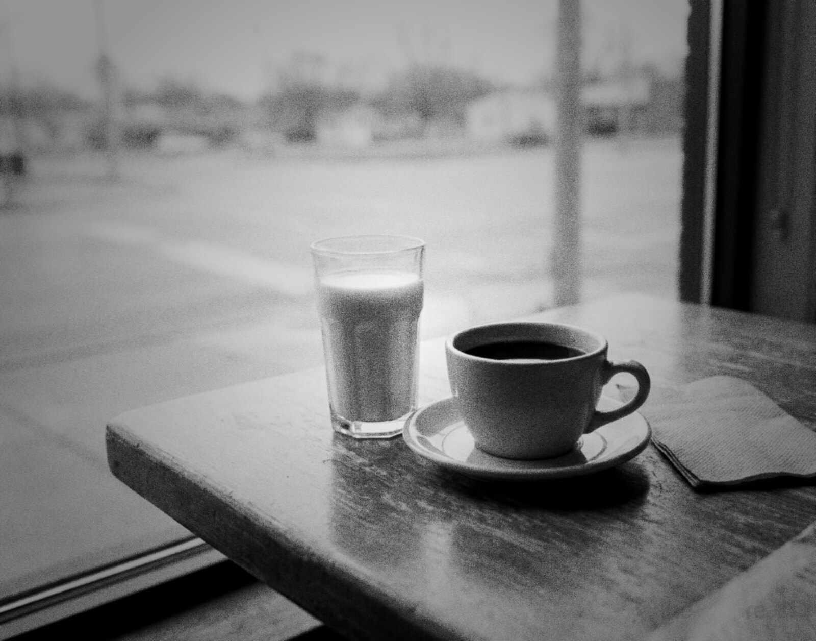A glass of milk and a cup of black coffee on a saucer on a worn wooden cafe table by a rainy street window, black and white photograph © The Voice Realm