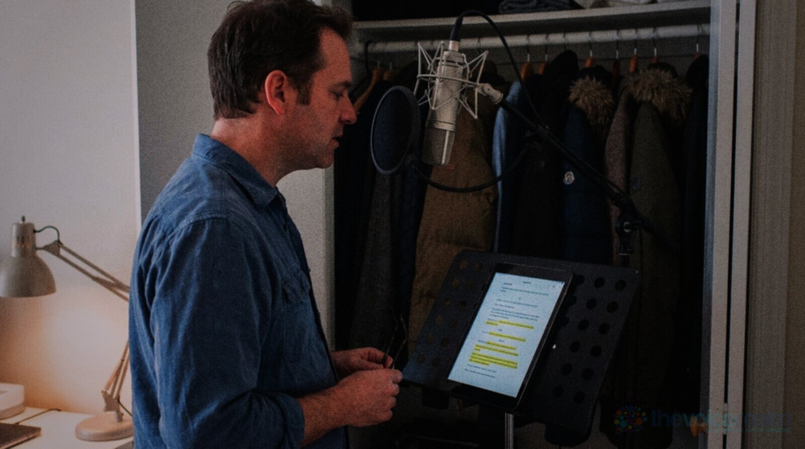 A man in a blue shirt recording in a converted wardrobe at home, standing slightly to one side of a condenser microphone that is placed above his mouth and angled down toward the corner of his lips through a round pop filter, a tablet script on a music stand, coats behind him © The Voice Realm