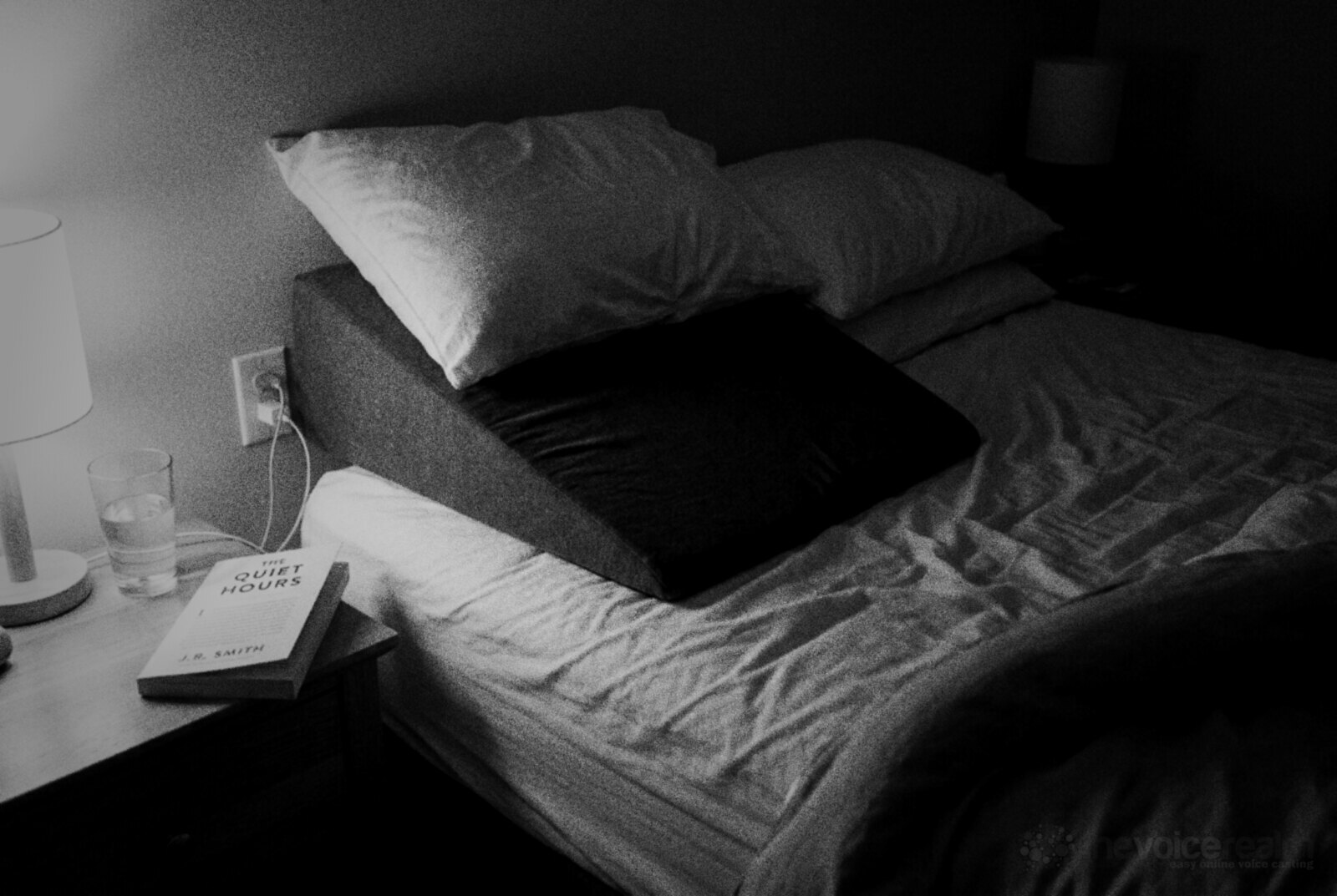 A bed at night with a foam wedge raising the head end under two pillows, a lamp, a glass of water and a paperback on the bedside table, black and white photograph © The Voice Realm