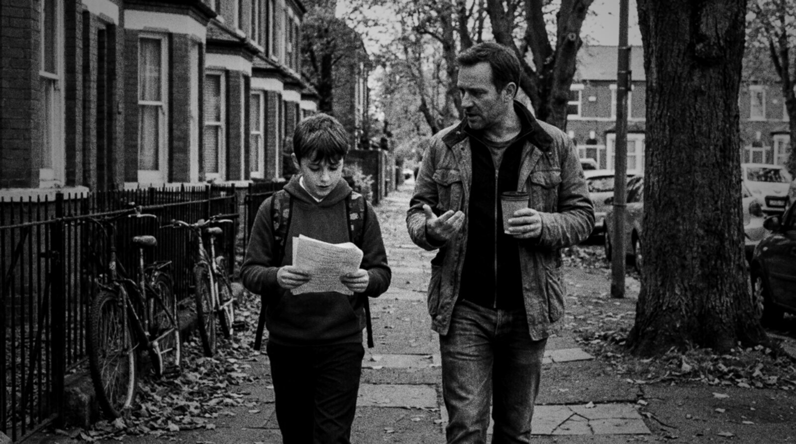 A father with a takeaway coffee talks with his son of about eleven, who reads printed script pages as they walk down a leafy street of brick terraced houses, black and white photograph © The Voice Realm