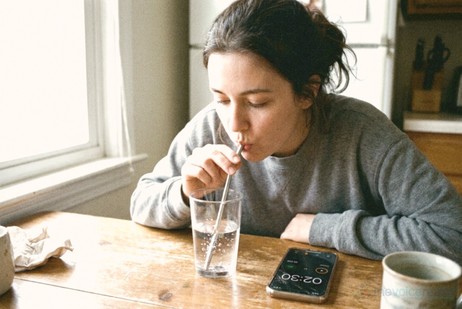 A young woman at a kitchen table hums through a metal straw into a glass of water, small bubbles rising, with a phone timer and a mug beside her, in bright phone snapshot colours © The Voice Realm