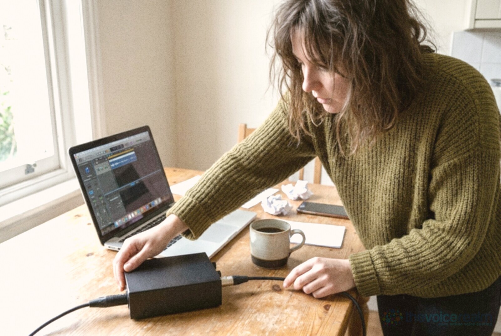 A woman in an olive knitted jumper pushes an XLR cable into a small black audio interface on a wooden kitchen table, an open laptop and a mug of coffee beside her in daylight from the window, in bright everyday phone photo colours © The Voice Realm