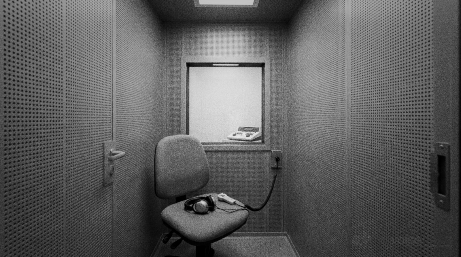 Black and white photo of an empty hearing test booth with perforated metal walls, a chair holding audiometer headphones and a response button, and an audiometer visible through the observation window © The Voice Realm