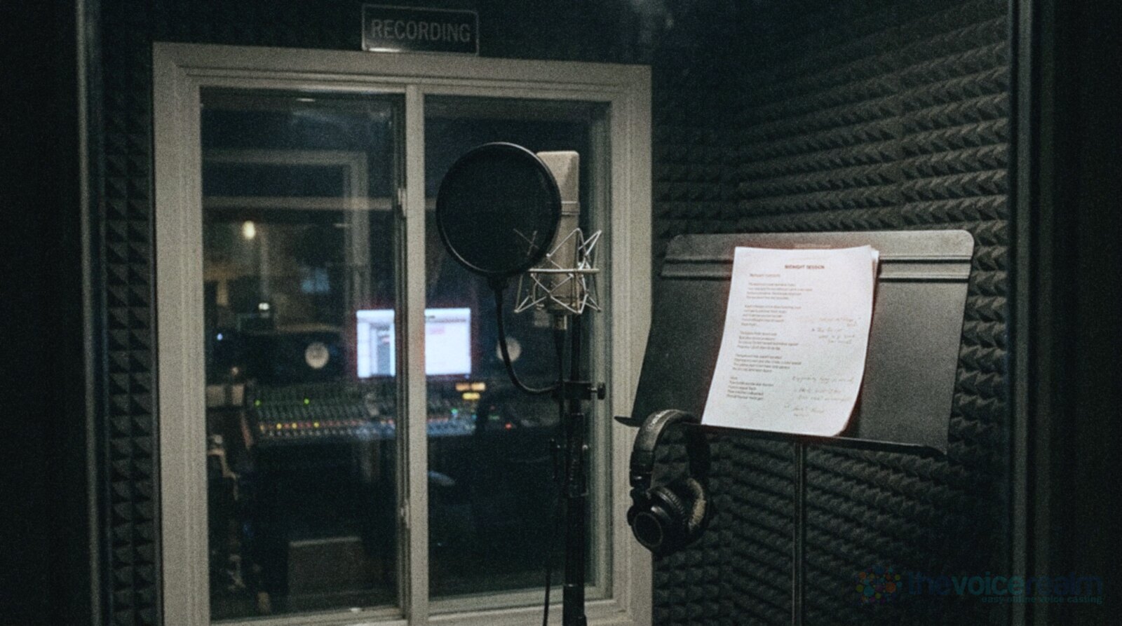 An empty recording booth at night with a large studio microphone behind a pop filter, a music stand holding a lyric sheet, headphones hanging from the stand and a control room glowing through the window © The Voice Realm