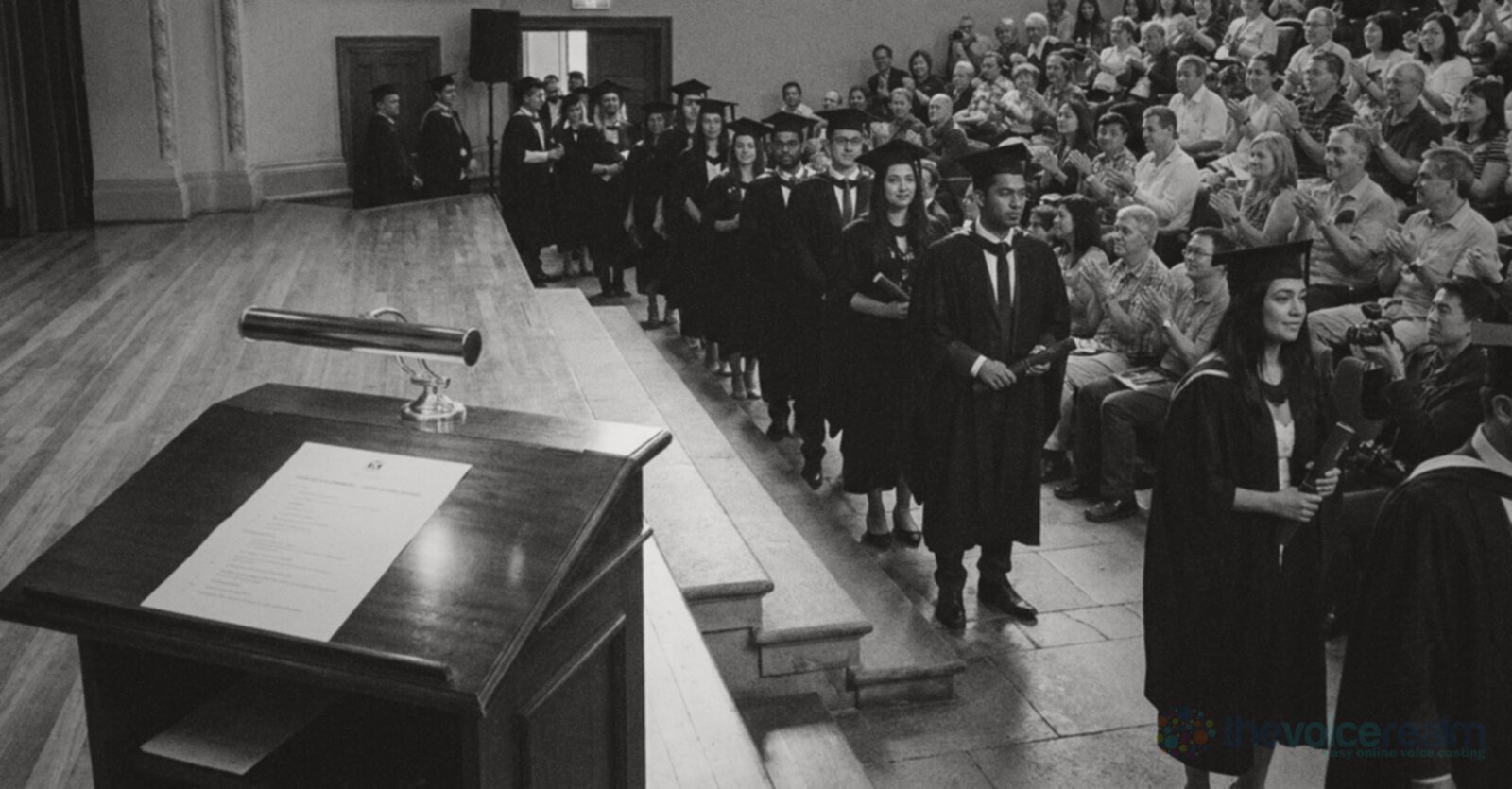 Graduates in black gowns and mortarboards queue at the foot of the stage in a grand university hall while families applaud, a lectern with a printed list of names in the foreground © The Voice Realm