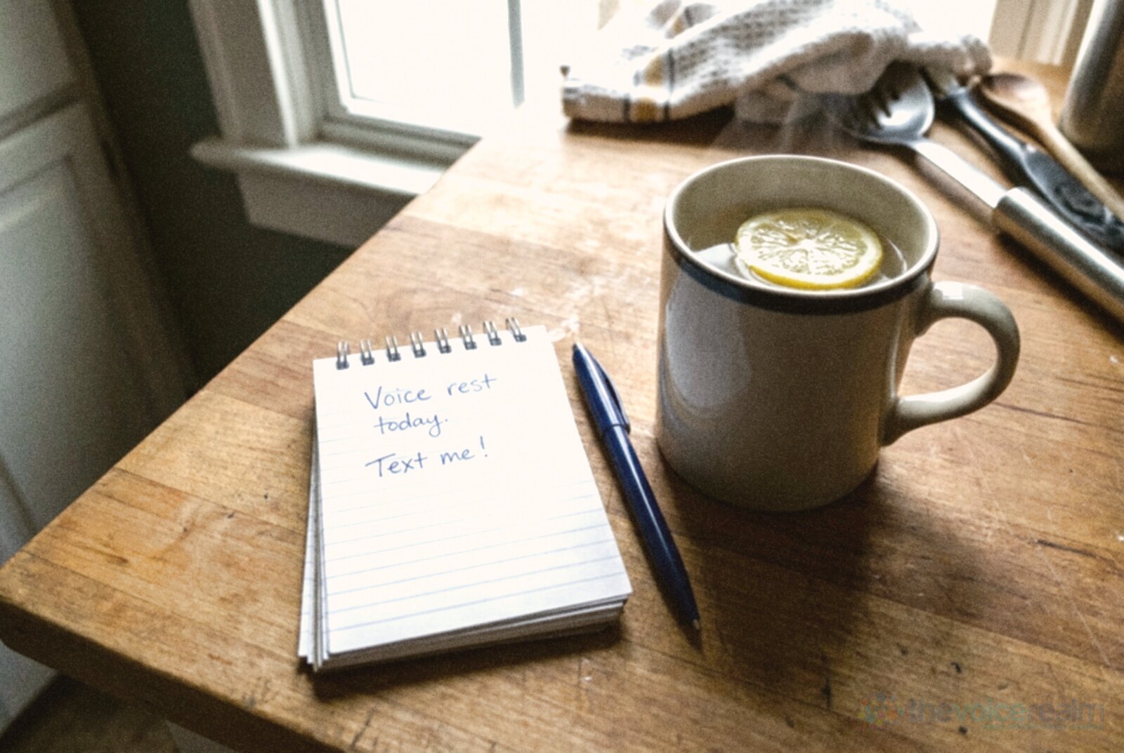 A small spiral notepad on a wooden kitchen counter with Voice rest today. Text me! handwritten in blue ink, a pen and a mug of warm water with a slice of lemon © The Voice Realm