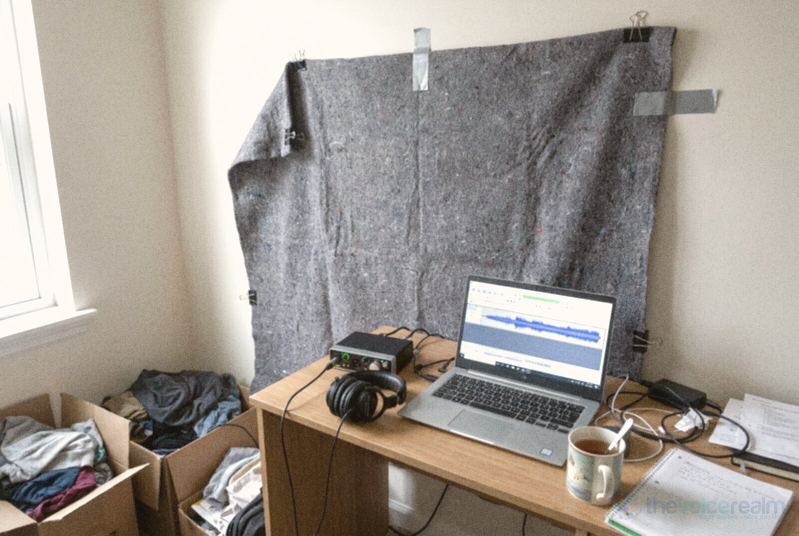 A spare bedroom set up for recording voice: a grey moving blanket taped to the wall, a small audio interface, closed-back headphones and a laptop showing a waveform on a desk, with a mug of tea and boxes of clothes © The Voice Realm