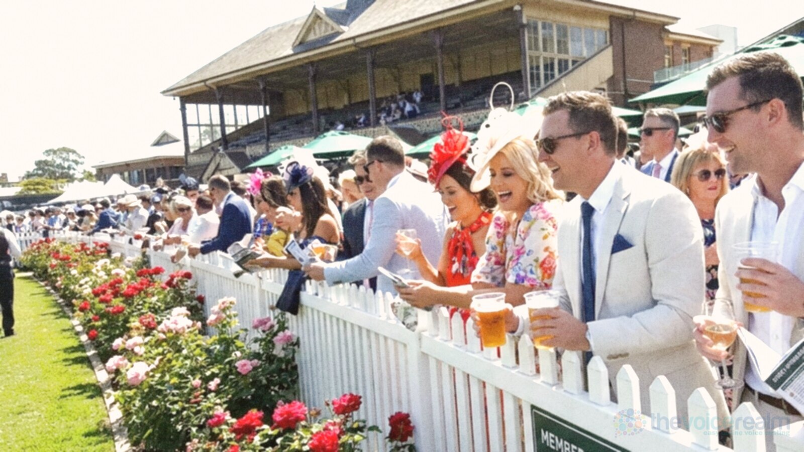 Racegoers in light suits, bright dresses and fascinators leaning on a white rail beside rose beds in front of a grandstand at an Australian racecourse on a sunny spring day © The Voice Realm