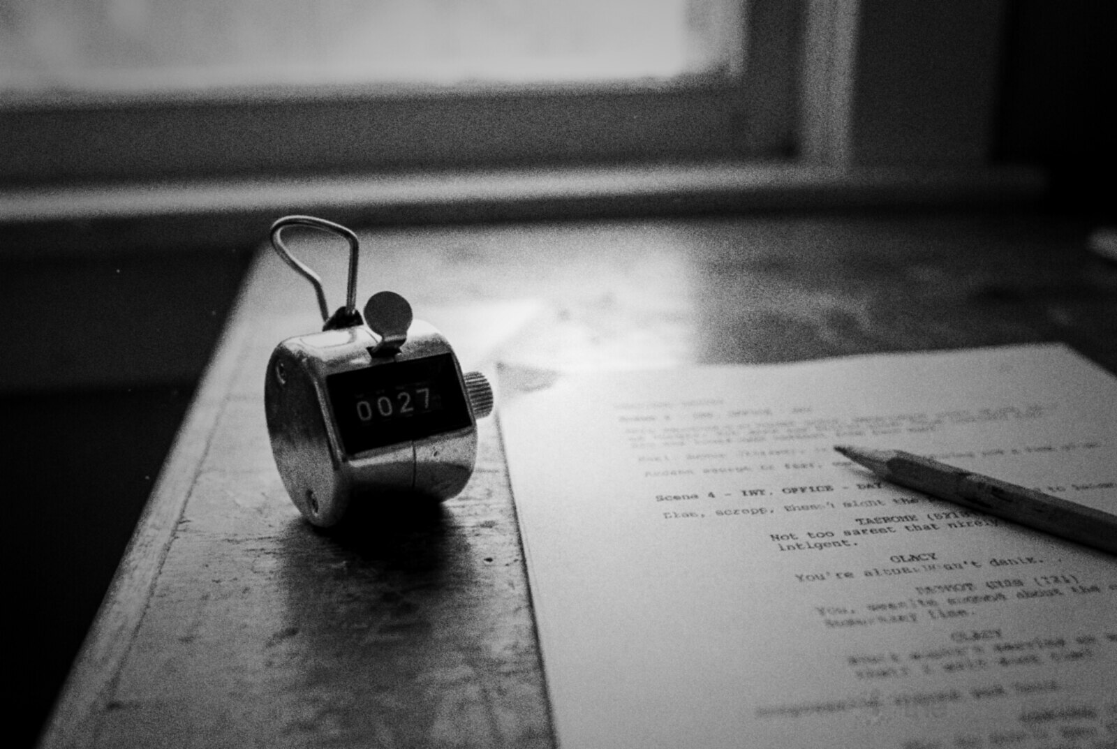 Black and white photo of a silver hand tally counter reading 0027 on a wooden desk by a window, beside a printed script page and a pencil © The Voice Realm