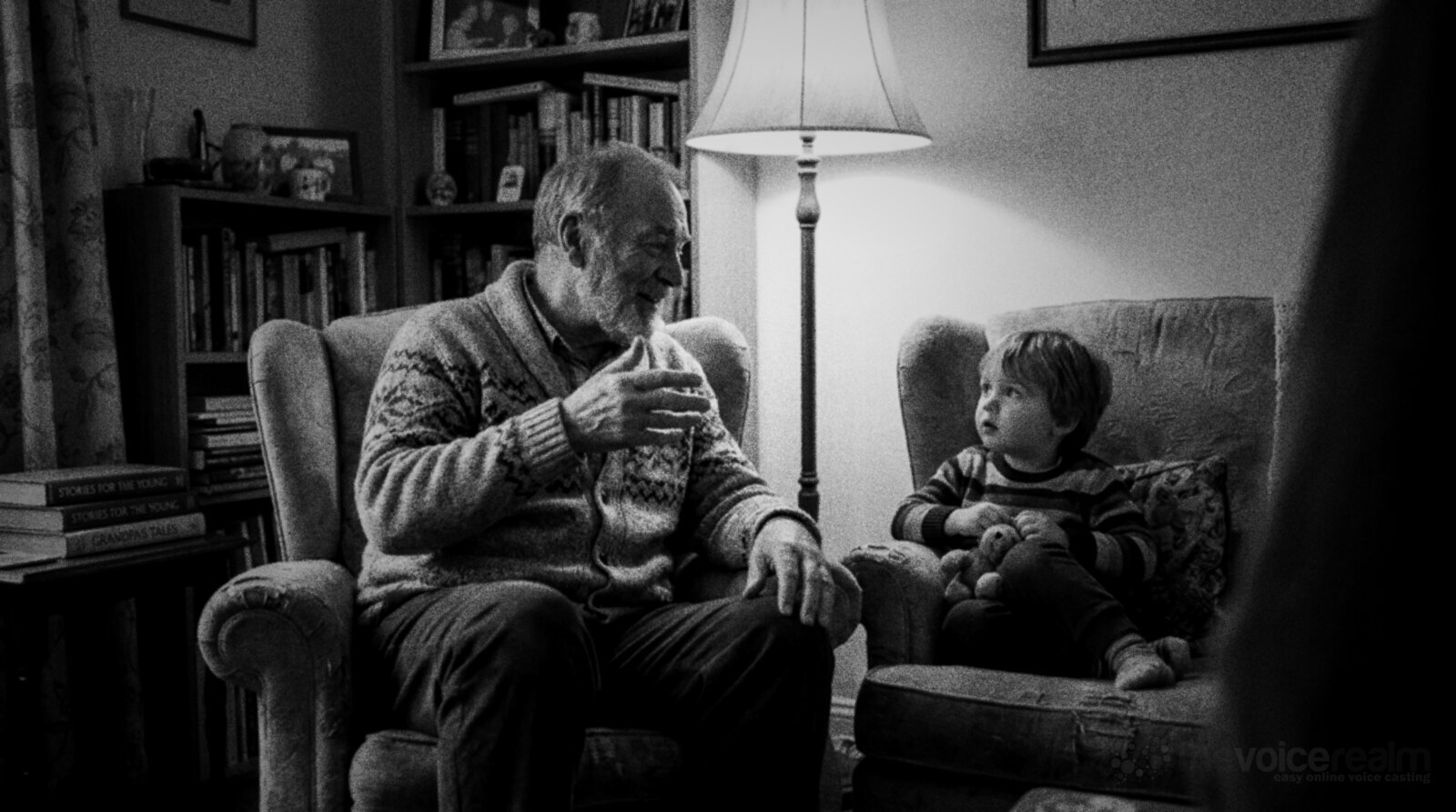 An older man in a cardigan telling a story to a small child in a lamplit living room, black and white photograph © The Voice Realm