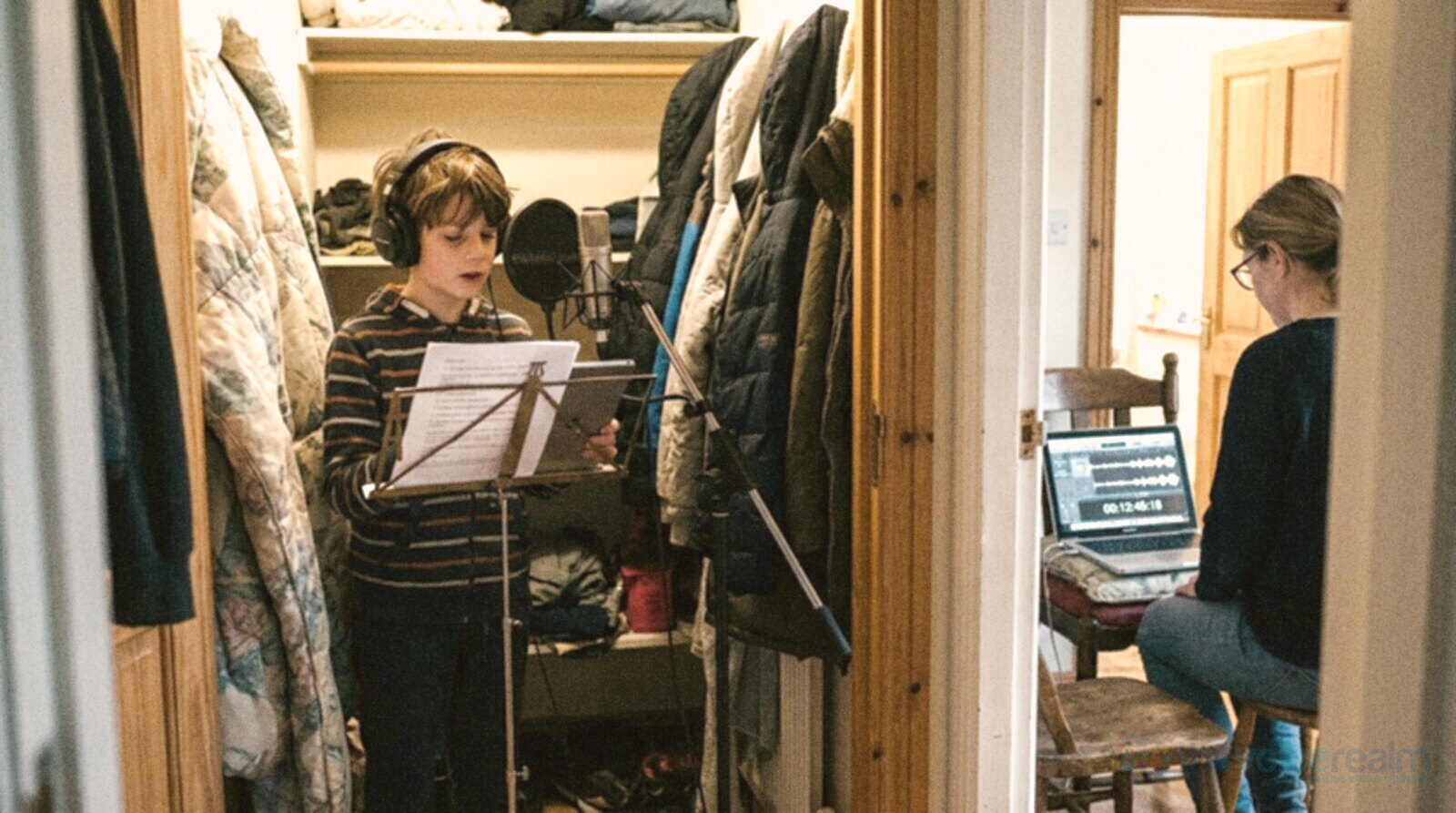 A boy in headphones reads from a music stand at a microphone with a pop filter inside a wardrobe lined with coats and a duvet, while his mother watches the recording on a laptop just outside the door © The Voice Realm