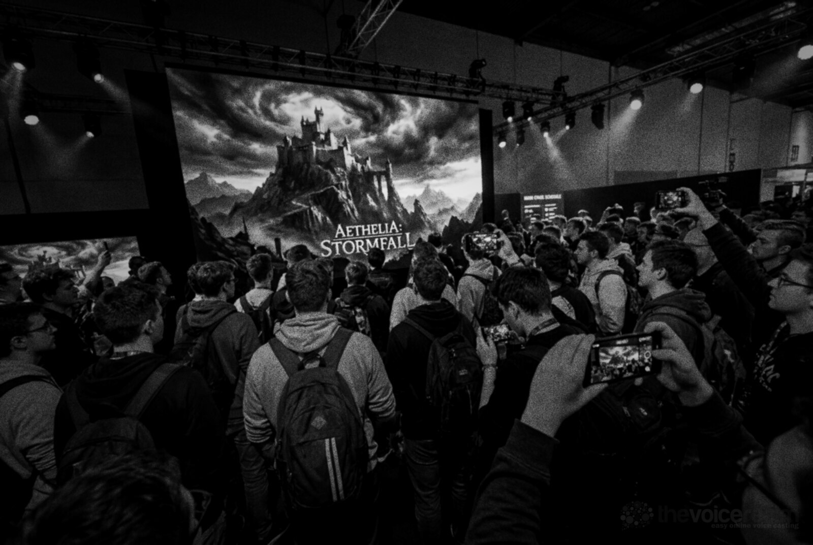 A crowd at a video game expo watching a huge screen showing a fantasy castle on a mountain under storm clouds, many holding up phones to film it, black and white photograph © The Voice Realm
