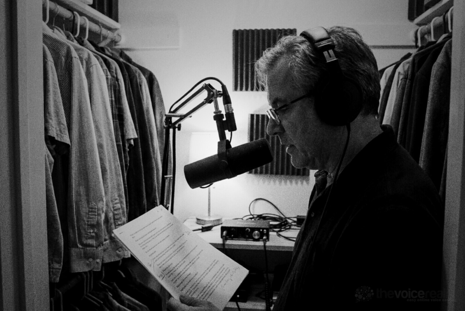 A grey-haired man in glasses and closed-back headphones reads a script into a dynamic broadcast microphone on a boom arm inside a walk-in clothes closet with a few foam panels, a small red audio interface on the shelf beside him, black and white photograph © The Voice Realm