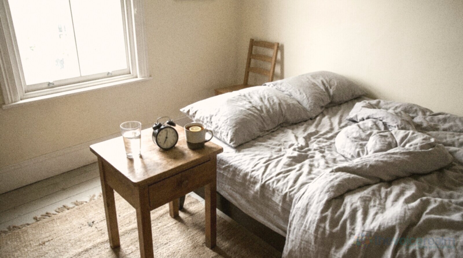 An empty bedroom in early morning light with a rumpled linen bed and a wooden bedside table holding a glass of water, a small alarm clock and a mug with a slice of lemon, in bright, flat phone photo colours © The Voice Realm