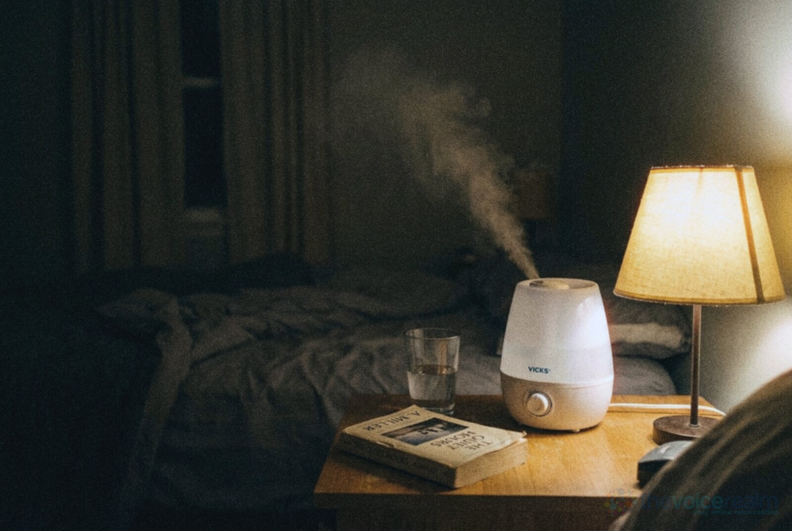 A small white humidifier sending up a plume of mist on a bedside table at night, beside a glass of water, a paperback and a warm lamp © The Voice Realm