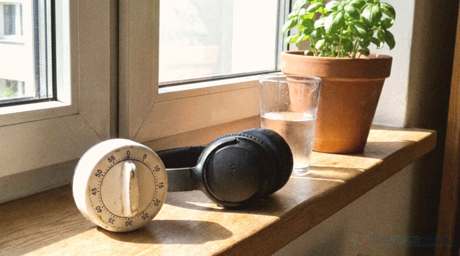 A white mechanical kitchen timer on a sunny windowsill beside folded headphones, a glass of water and a potted basil plant © The Voice Realm