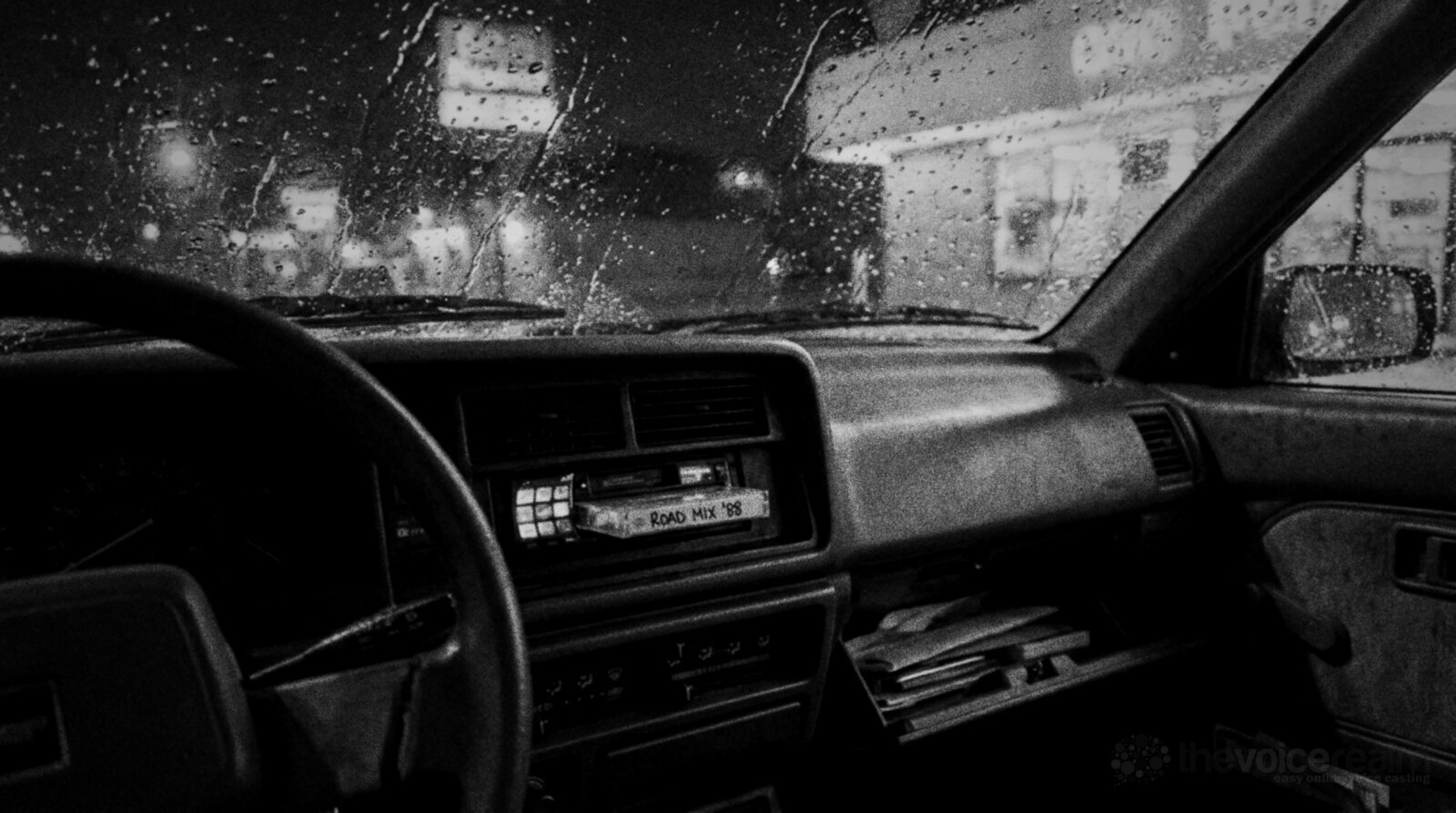 Black and white photo inside a parked 1980s car at night, a cassette labelled Road Mix '88 half pushed into the glowing dashboard stereo, rain on the windscreen © The Voice Realm