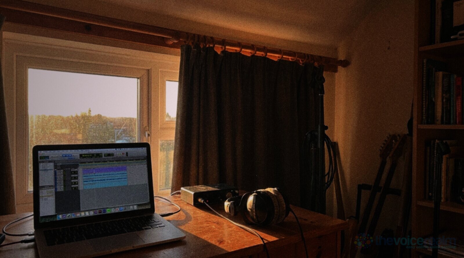 A spare room recording desk in low late afternoon sun, a laptop showing a recording session, an audio interface and headphones by a window with heavy curtains © The Voice Realm