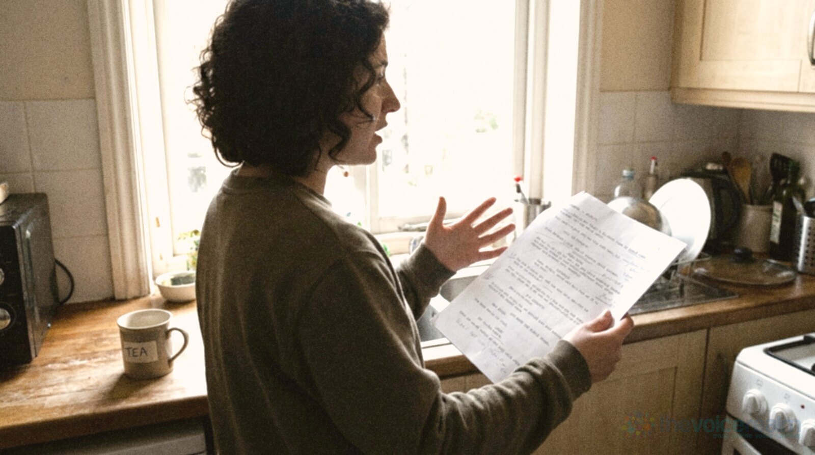 A person with dark curly hair stands in a bright home kitchen reading aloud from a printed script page, one hand mid gesture, a mug on the counter © The Voice Realm