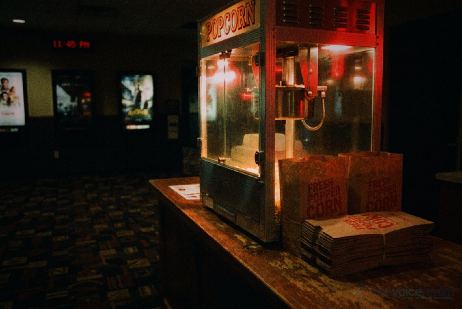 A glowing old-fashioned popcorn machine on the counter of a dim cinema lobby late at night, with paper popcorn bags stacked beside it and film posters lit on the far wall © The Voice Realm