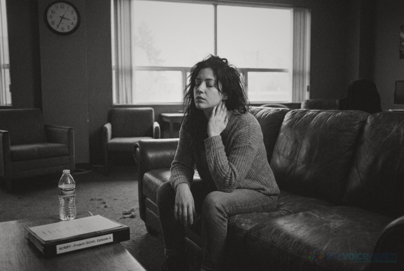 A tired woman rests on a worn leather sofa in a studio lounge with her eyes closed and a hand on her throat, a water bottle and a script binder on the table in front of her, black and white photograph © The Voice Realm
