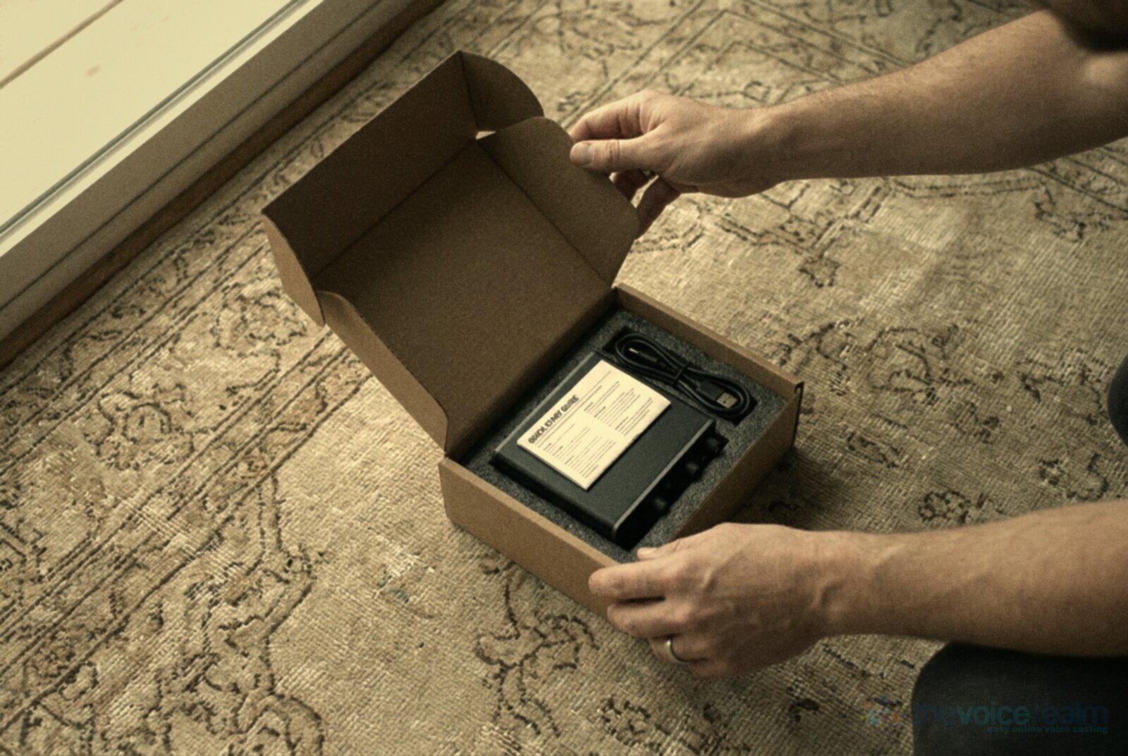 Seen from above, two hands open a small cardboard box on a patterned rug to reveal a compact black audio interface in a foam tray with a coiled USB-C cable and a quick start guide, in soft faded warm film colours © The Voice Realm
