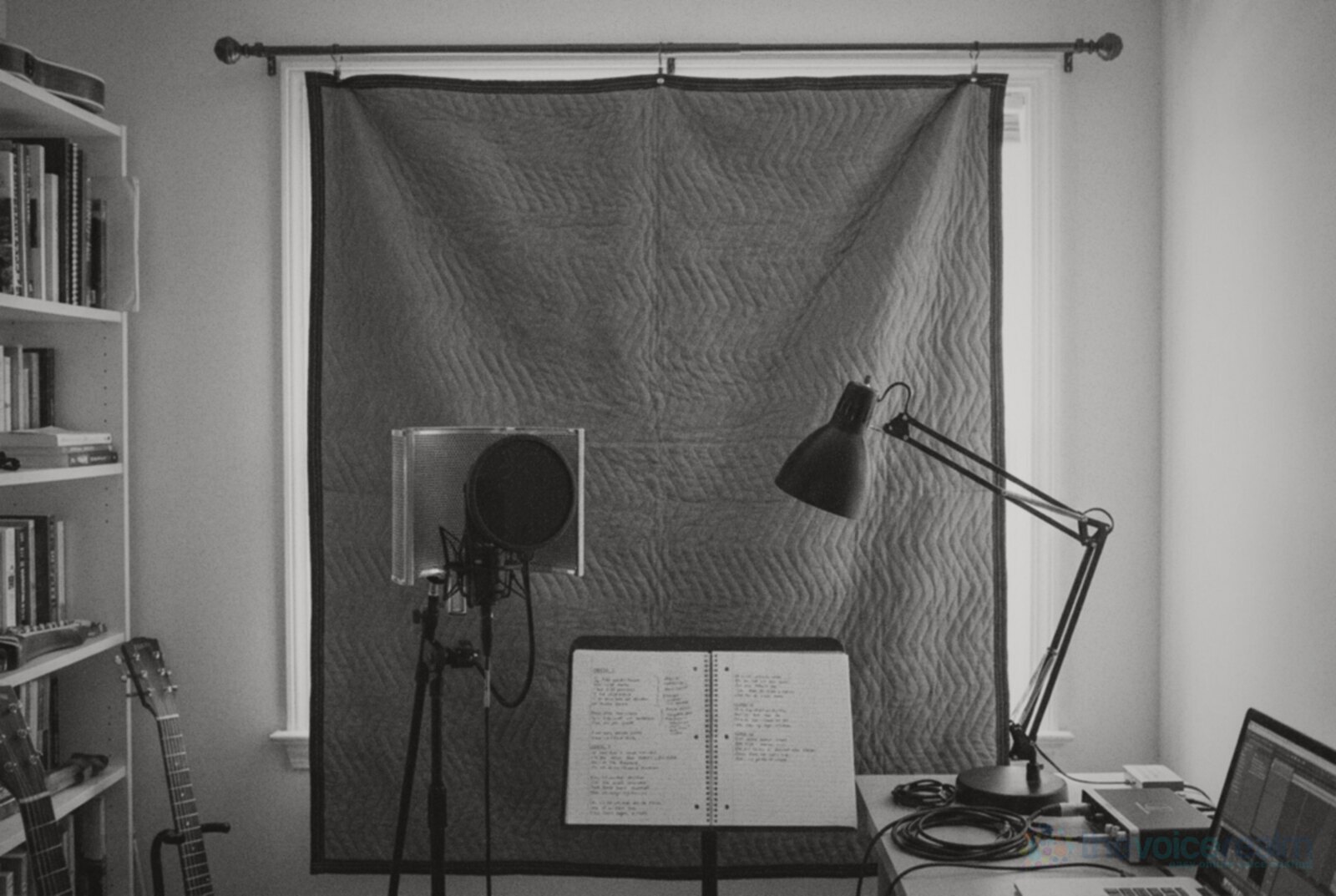 A home recording corner in a spare room with a grey quilted moving blanket hung from a curtain rod over the window, a microphone with a pop filter and reflection shield, a music stand with a notebook and a desk lamp, black and white photograph © The Voice Realm