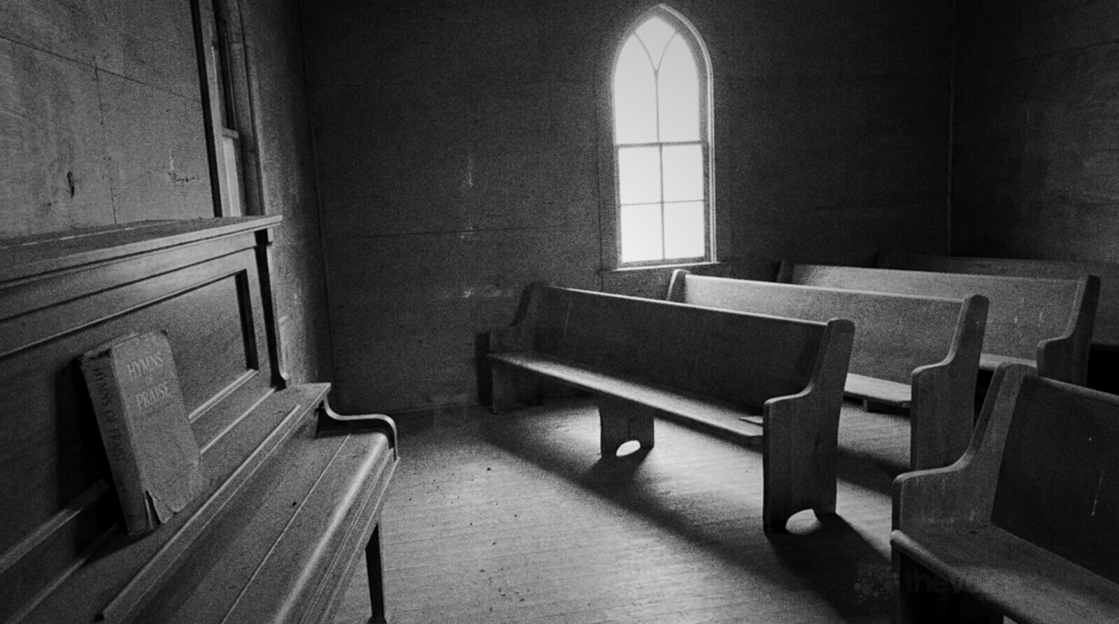 An empty wooden church hall with rows of plain pews, a tall arched window and an old upright piano with a hymn book against the wall, black and white photograph © The Voice Realm