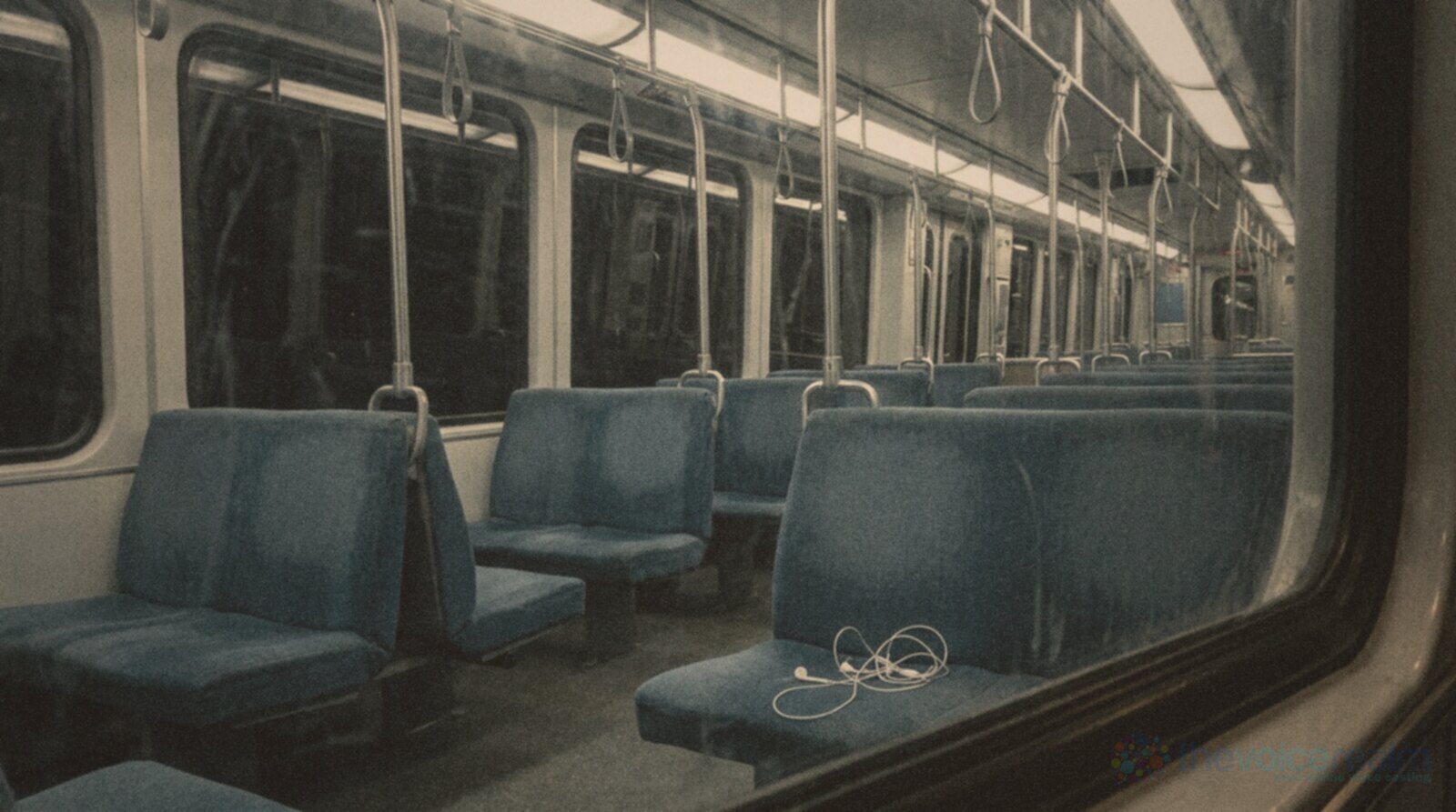 An empty commuter train carriage at night with rows of blue seats and grab poles, a pair of white wired earbuds left tangled on one seat © The Voice Realm