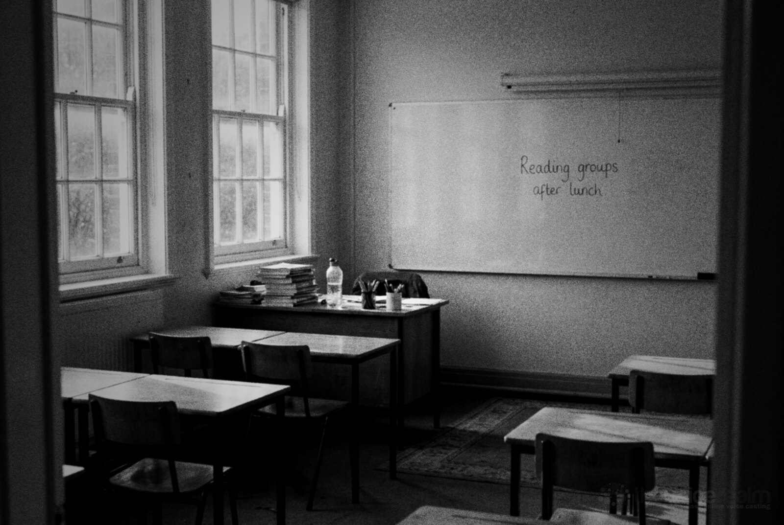 Black and white photo of an empty classroom after the bell, chairs pushed in under the desks and a whiteboard reading Reading groups after lunch © The Voice Realm