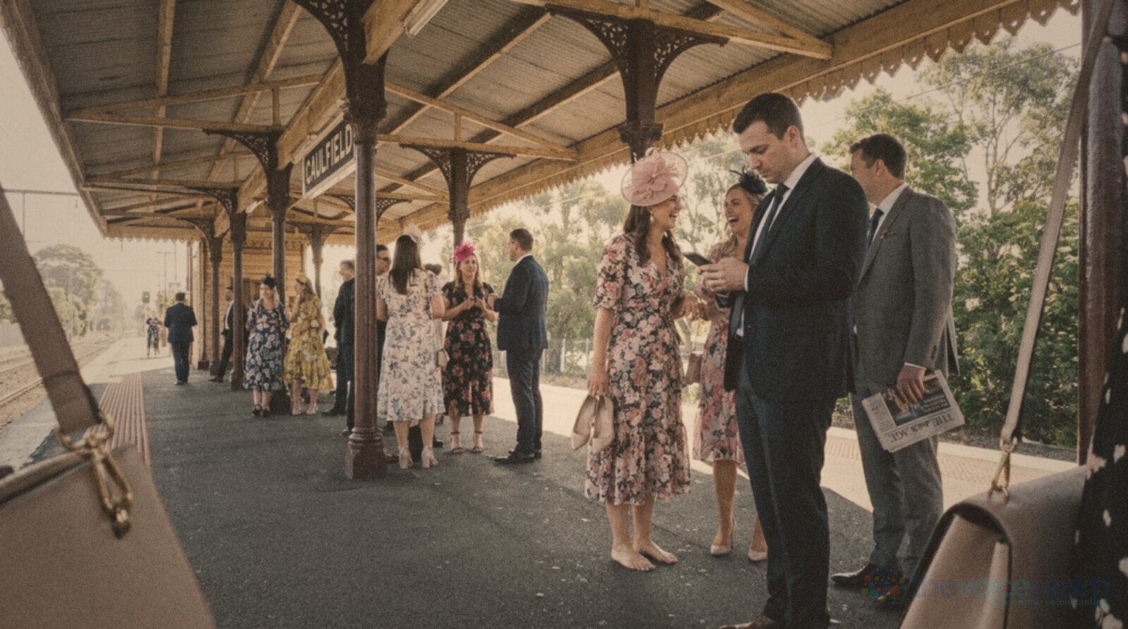 Racegoers in floral dresses, suits and fascinators waiting under the iron canopy of a suburban Melbourne railway platform on a spring morning, one woman holding her heels © The Voice Realm