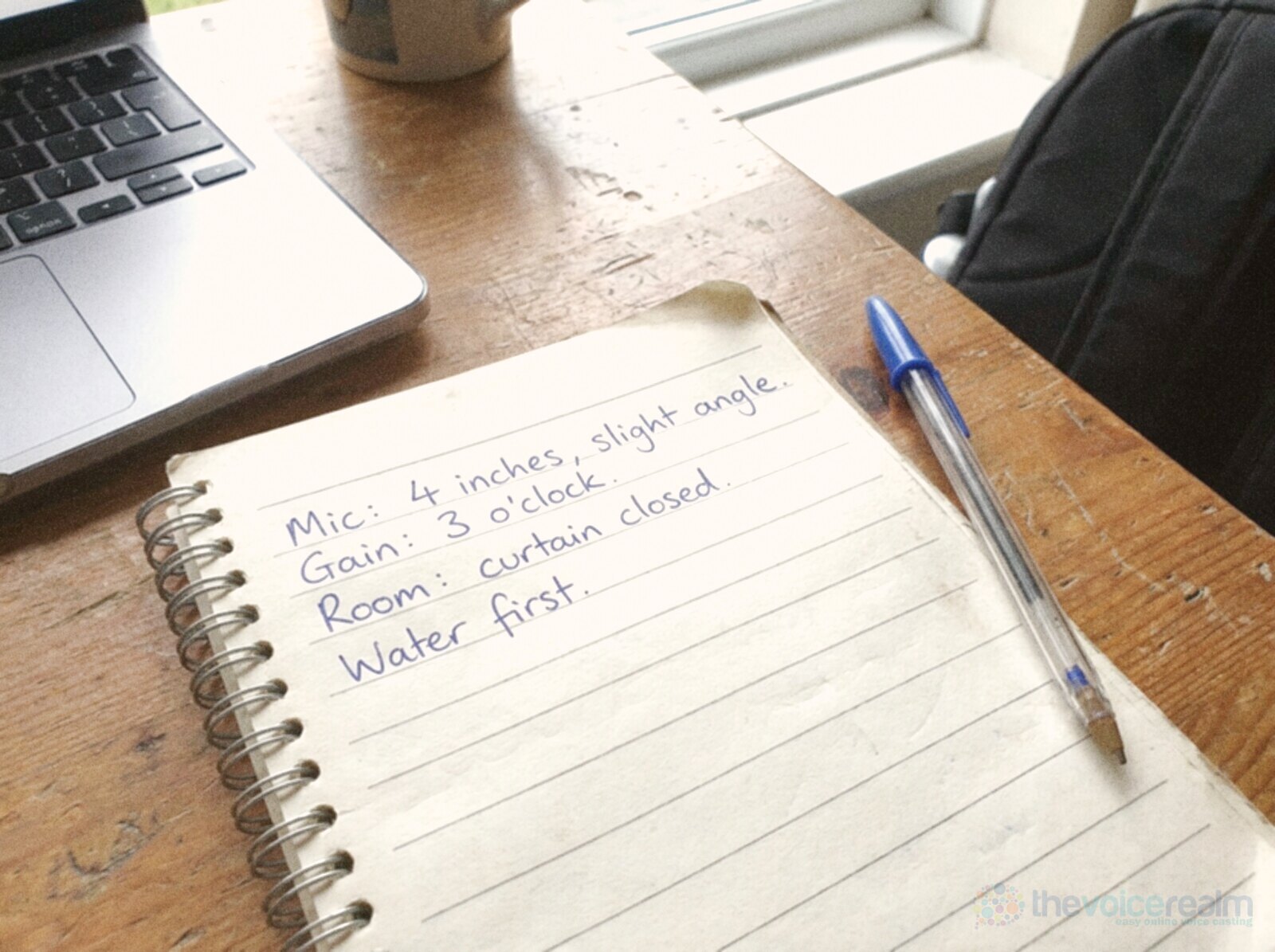 A spiral notebook open beside a laptop with handwritten session notes: mic 4 inches, slight angle, gain 3 o'clock, room curtain closed, water first © The Voice Realm