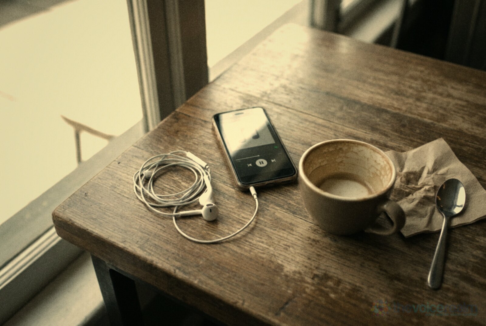 White wired earbuds coiled on a cafe table beside a phone playing audio, an empty coffee cup and a crumpled napkin © The Voice Realm