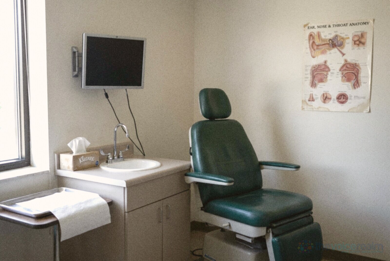 An empty ear, nose and throat clinic room in daylight with a green patient chair, a wall monitor, a small sink, a box of tissues and an anatomy poster © The Voice Realm