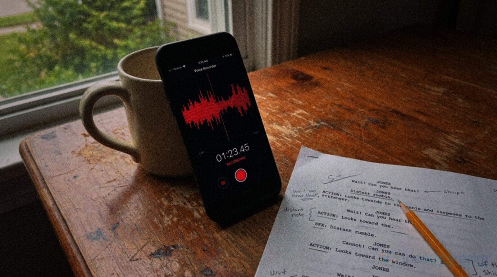 A smartphone leaning against a mug on a worn wooden desk, its voice recorder app showing a red waveform, beside a script page marked with pencil © The Voice Realm