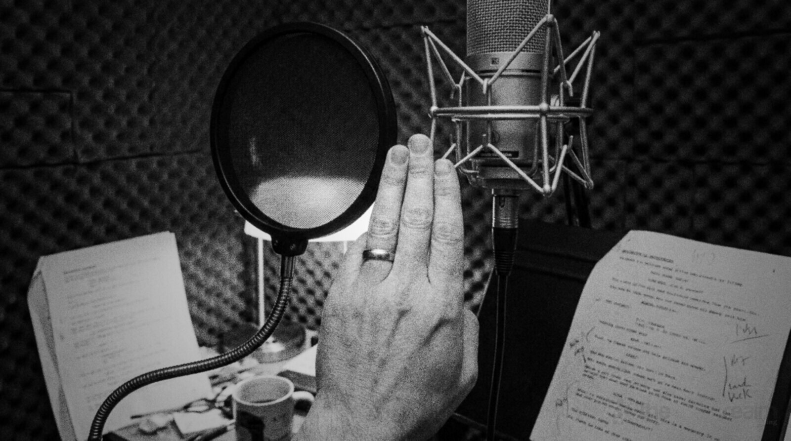 Black and white photo in a small foam lined home booth of a hand held sideways with four fingers filling the gap between a round pop filter on a gooseneck and a large diaphragm condenser microphone in a shock mount, scripts on a stand behind © The Voice Realm