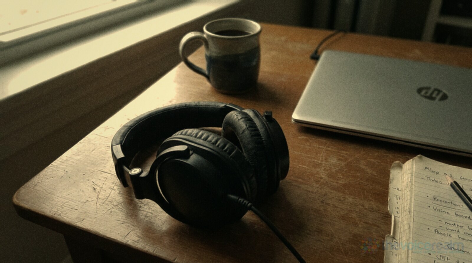A pair of black closed-back over-ear headphones resting on a scratched wooden desk beside a silver laptop, a ceramic mug and a notebook, in soft window light © The Voice Realm