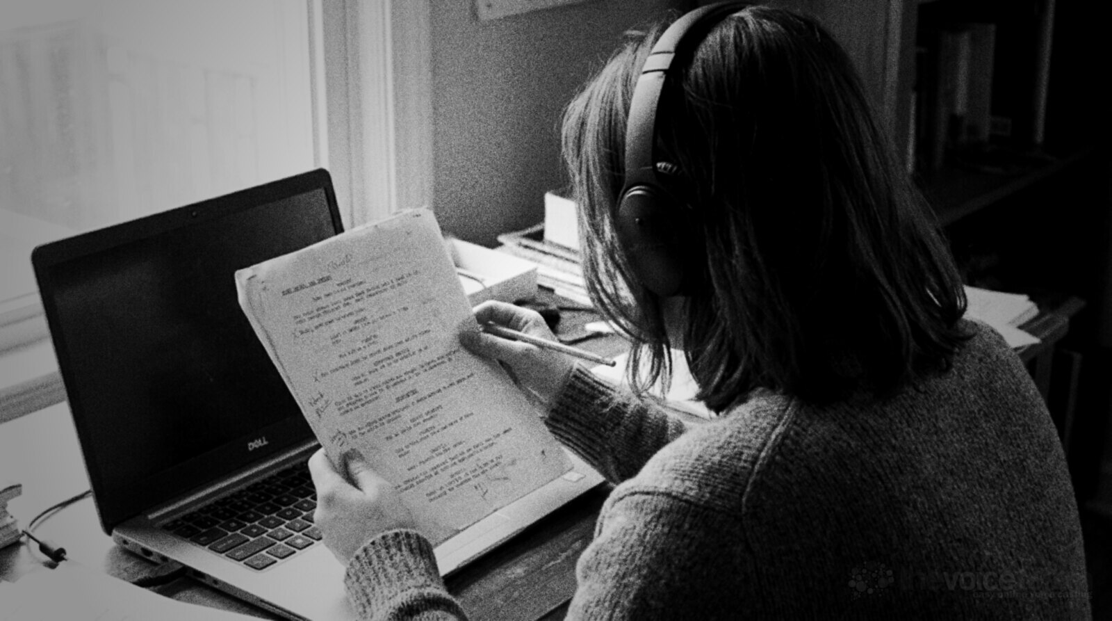 Black and white photo of a person seen from behind wearing closed-back headphones at a desk, reading a marked-up script page with a pencil in front of a laptop © The Voice Realm