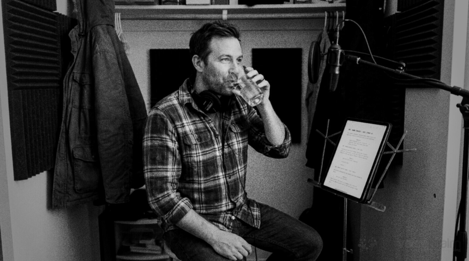 Black and white photo of a man in a plaid shirt with headphones around his neck sipping a glass of water on a stool in a home recording closet lined with foam panels, a tablet script on a music stand beside him © The Voice Realm