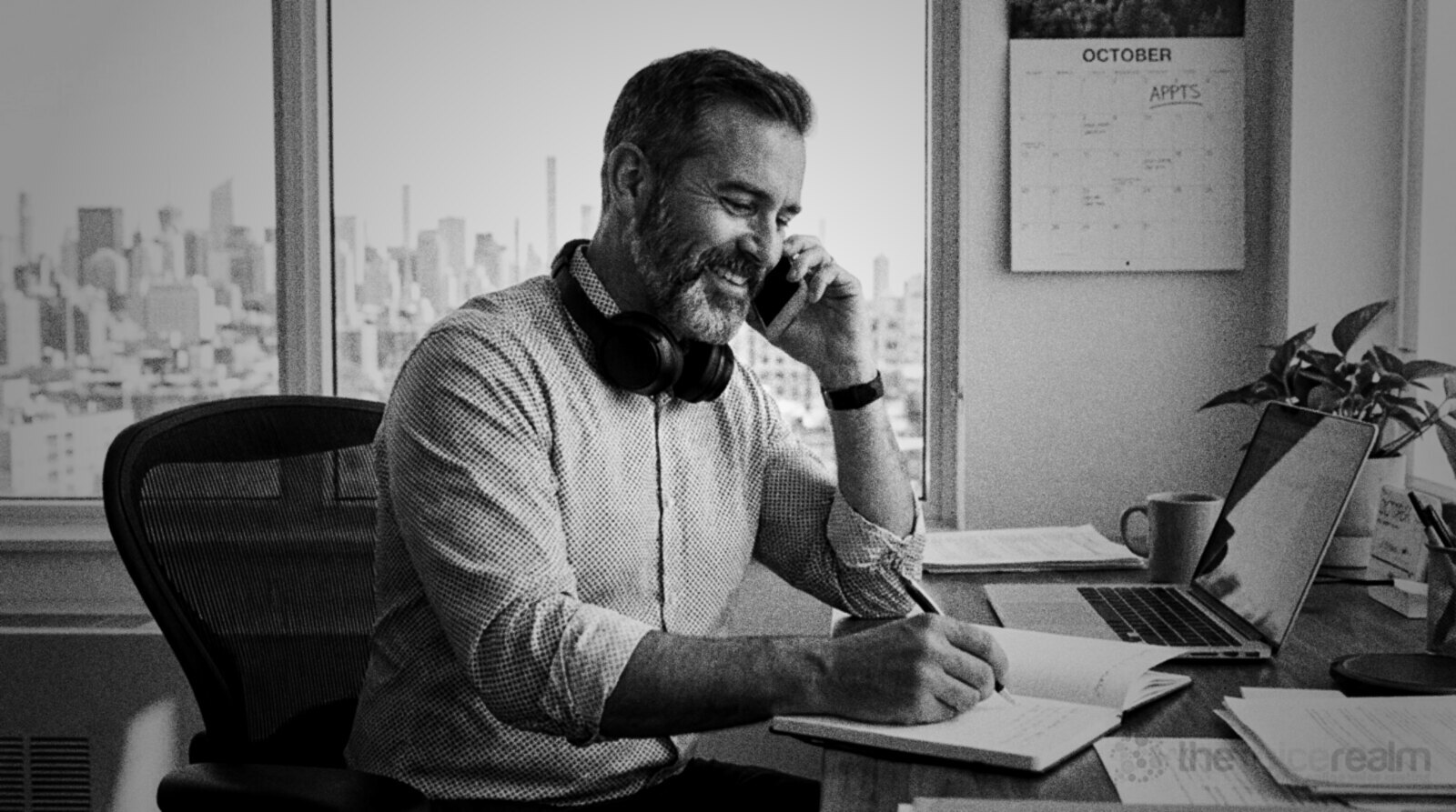 A smiling man on a phone call at a home office desk, writing in a notebook, headphones around his neck and a city skyline through the window © The Voice Realm