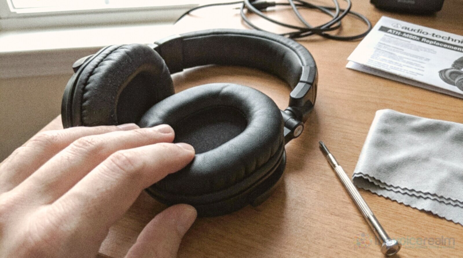 A hand pressing a soft replacement ear cushion onto a pair of black headphones on a desk, with a small screwdriver and a grey cloth beside them © The Voice Realm