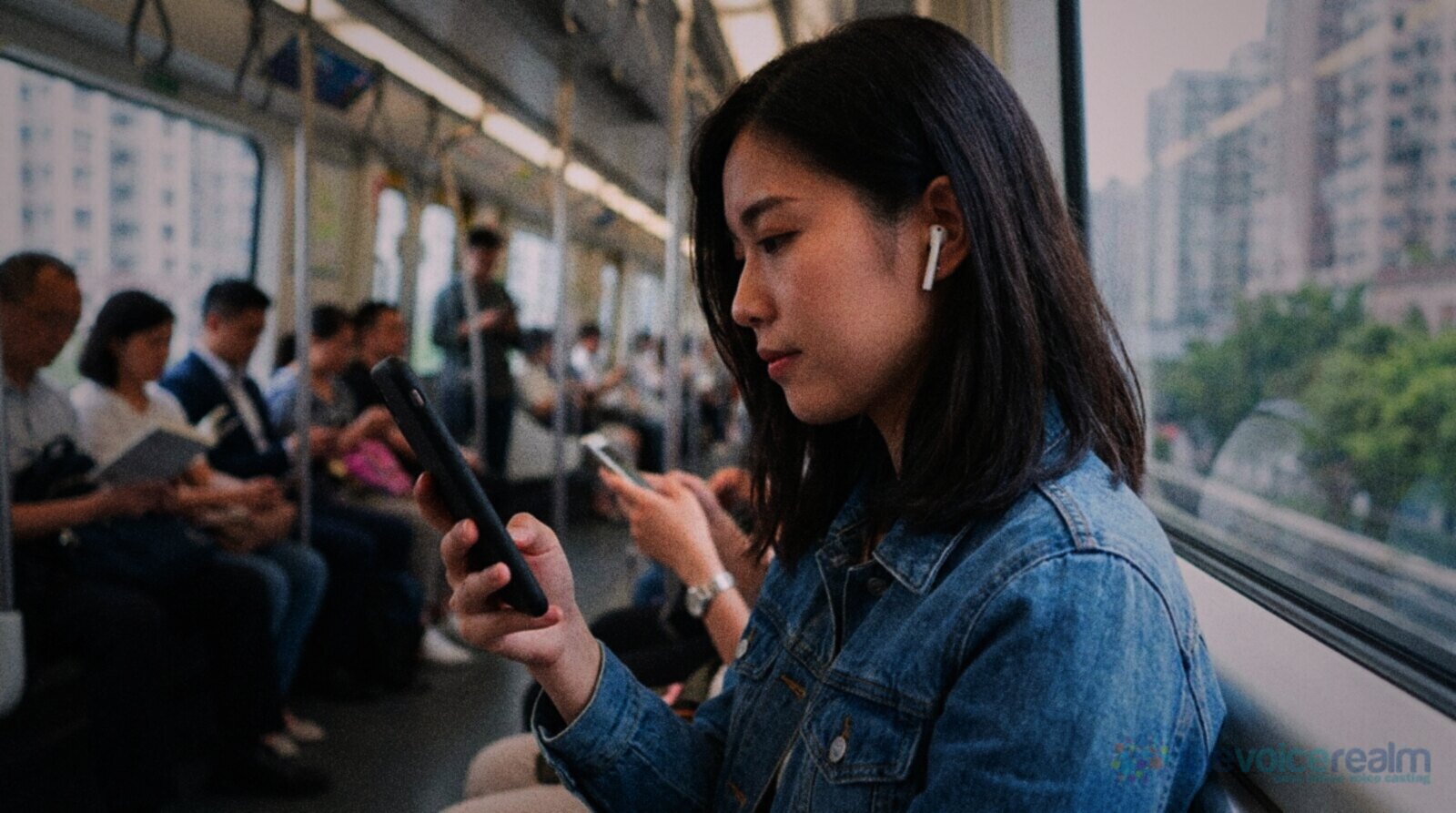A young woman in a denim jacket with wireless earbuds watching a video on her phone on a commuter train, other passengers behind her © The Voice Realm