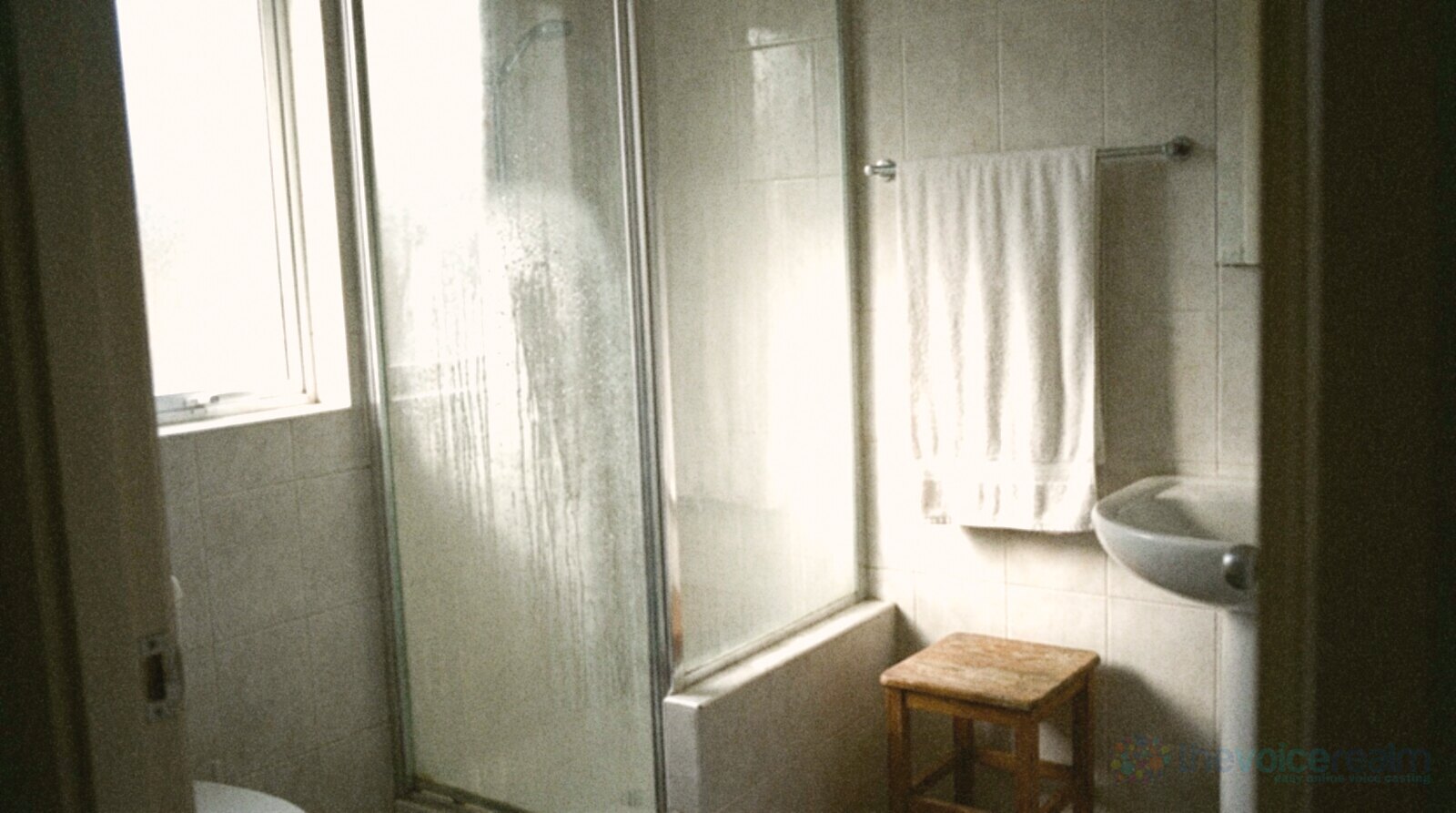 A small tiled home bathroom with the hot shower running behind a fogged glass screen, droplets running down the glass and a white towel on a rail © The Voice Realm