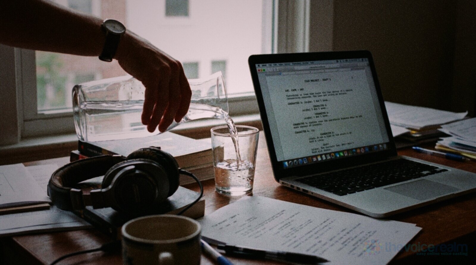 A hand pouring water from a glass jug into a tumbler on a wooden desk beside an open laptop showing a script, a pair of studio headphones and a mug © The Voice Realm